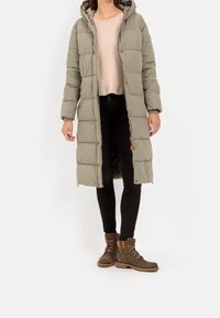 Długa, puchowa, oliwkowozielona zimowa kurtka z kapturem, noszona na jasnym swetrze i czarnych jeansach, zestawiona z brązowymi, sznurowanymi botkami za kostkę.