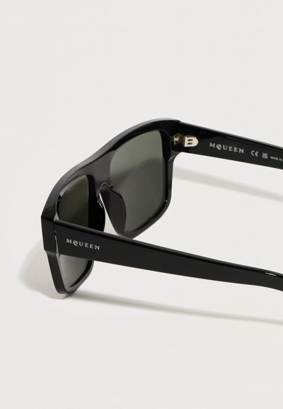 Μαύρα ορθογώνια γυαλιά ηλίου McQueen με σκουρόχρωμους φακούς, παχιούς γυαλιστερούς σκελετούς και ορατό όνομα εμπρόσθιας μάρκας στα πλαϊνά της βάσης.