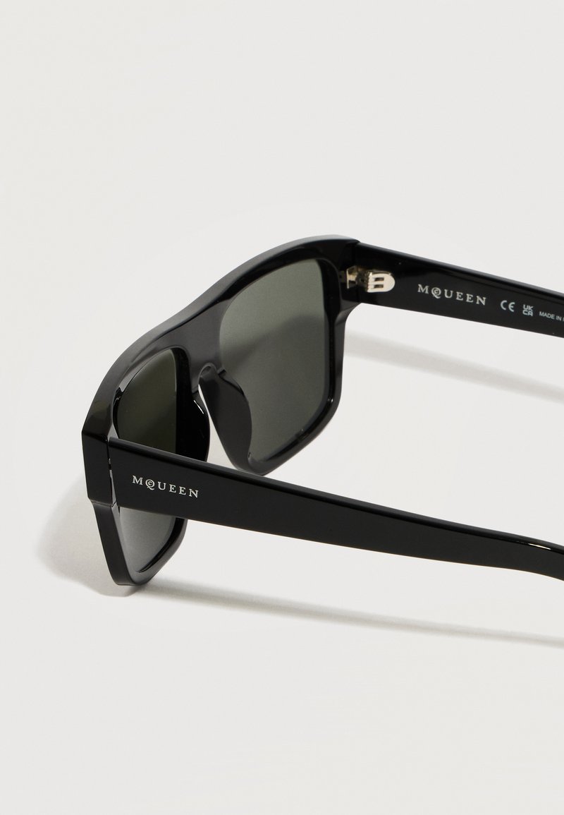 Lunettes de soleil McQueen rectangulaires noires avec verres foncés, montures épaisses et brillantes, et nom de la marque visible sur les branches.