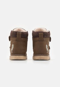 Bottines brunes à la cheville avec un dessus en laine, doublure duveteuse, bandes Velcro et semelle légère. Présente un motif de lion sur le côté extérieur.