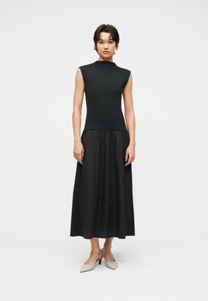 Kvinde stående iført en ærmeløs sort top med strukturerede skuldre og et langt sort rynket skjørt, kombineret med lyse beige spidse hæle.