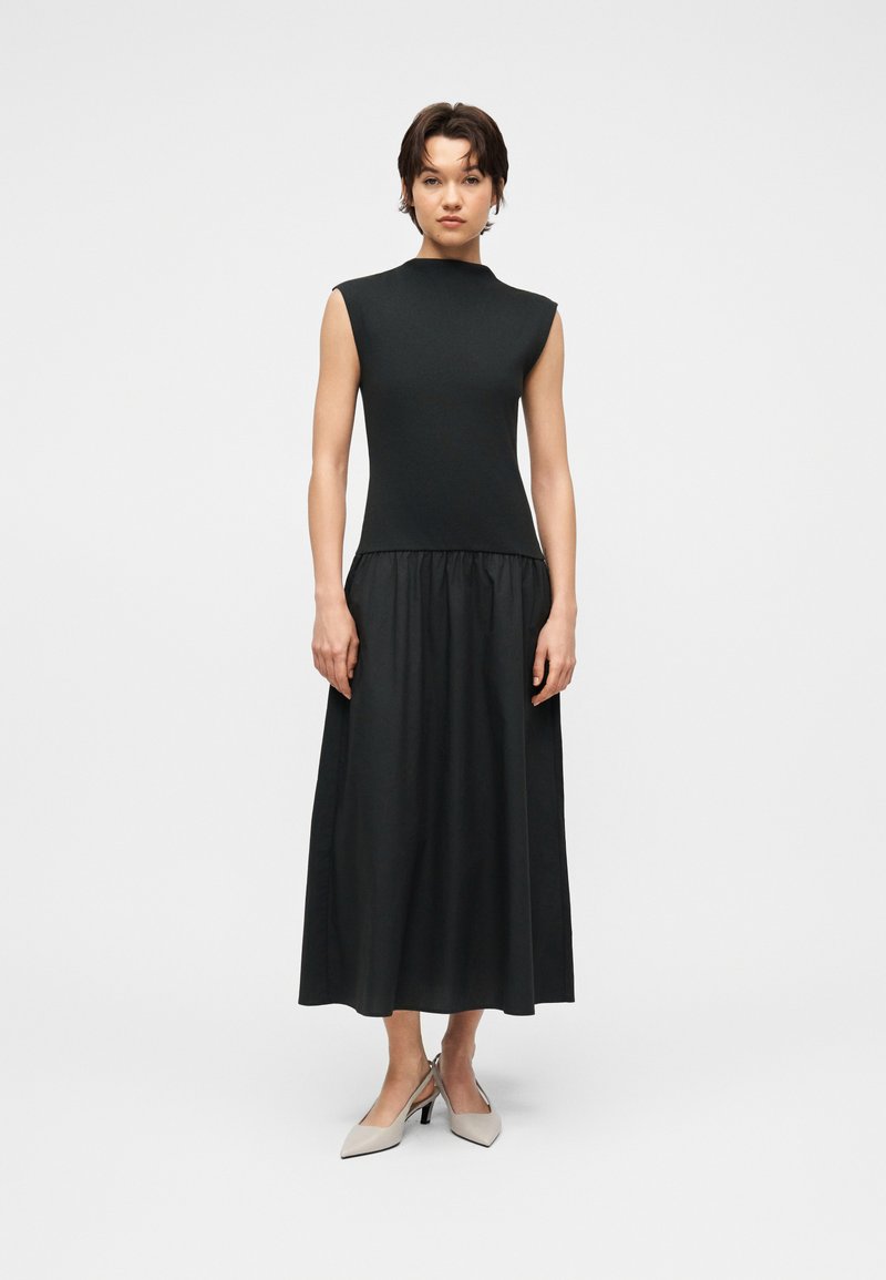 Femme debout portant un haut noir sans manches avec des épaules structurées et une longue jupe noire drapée, associée à des escarpins beiges clairs à bout pointu.