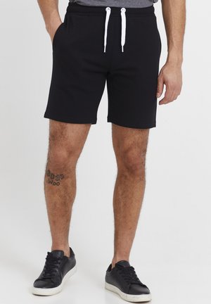 Pantalones cortos deportivos negros con cintura elástica y cordón, con acentos blancos, que cuentan con bolsillos laterales y un corte relajado, combinados con zapatillas negras.