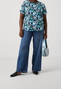 Haut à manches courtes floral en bleu, vert et noir, associé à un jean en denim bleu à jambes larges et des chaussures noires à enfiler, tenant un sac bleu clair.