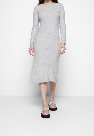 Femme portant une robe midi en maille côtelée light grey ajustée et des mocassins roses avec des sangles multicolores, se tenant devant un fond blanc uni.