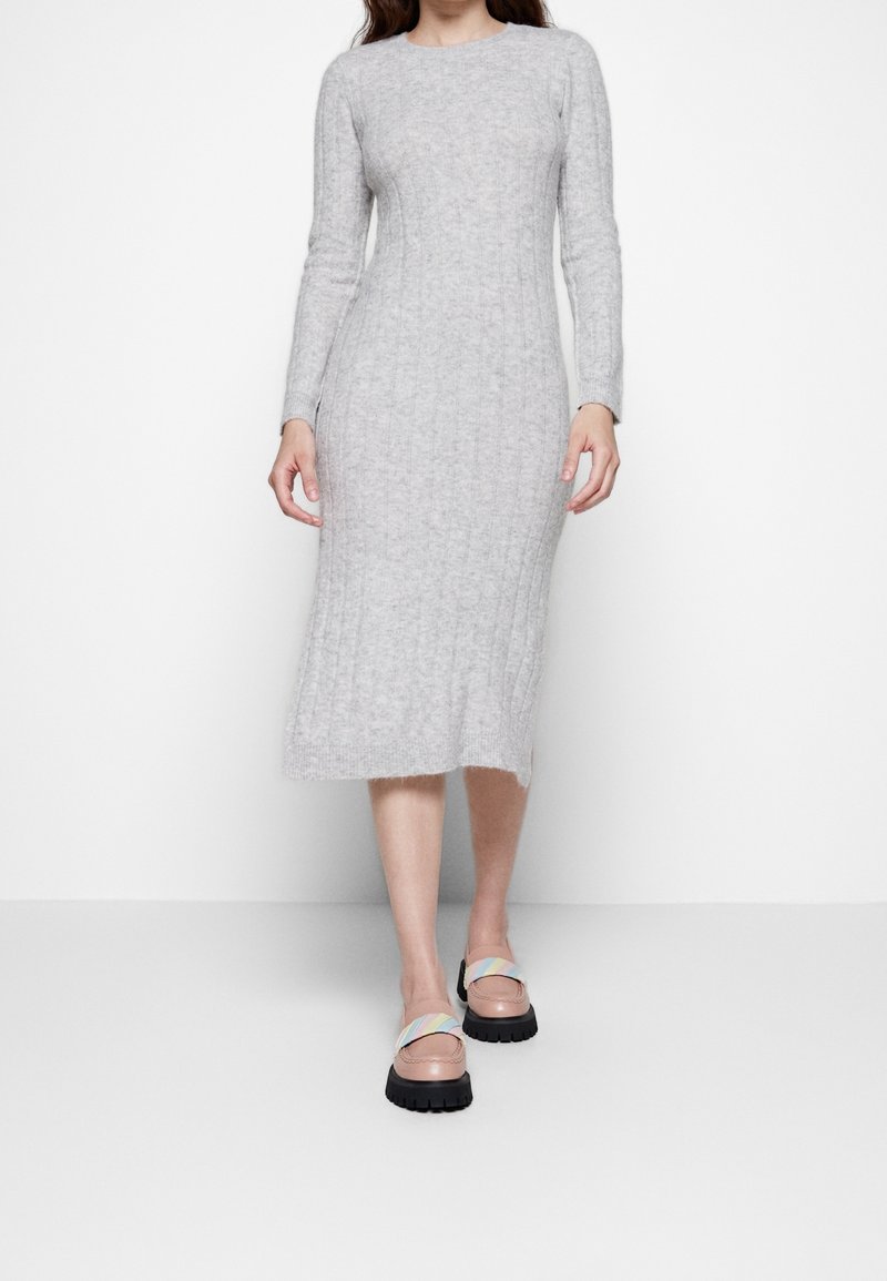 Donna che indossa un abito midi aderente grigio chiaro a costine e mocassini rosa con cinturini multicolori, in piedi su uno sfondo bianco semplice.