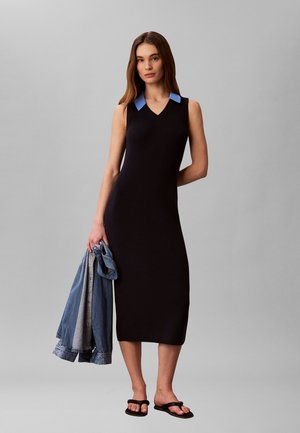 Kobieta w czarnej sukience midi bez rękawów z niebieskim kołnierzykiem, trzymająca kurtkę jeansową w jednej ręce i nosząca czarne sandały.