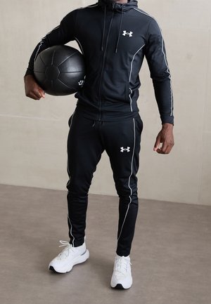 Osoba w czarnym sportowym dresie trzyma dużą czarną piłkę lekarską, ma na nogach białe buty sportowe, stoi na szarej podłodze.