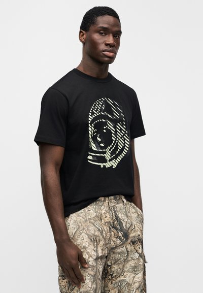 Μαύρο βαμβακερό T-shirt με έντονο, αφηρημένο γραφικό σε ανοιχτό πράσινο. Το μοντέλο φοράει μπεζ cargo παντελόνια με σχέδιο και κορδόνι στη μέση.