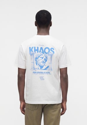 Homme portant un t-shirt blanc avec un motif bleu d'une tête de statue classique entre deux colonnes et le texte "KHAOS" dans le dos.