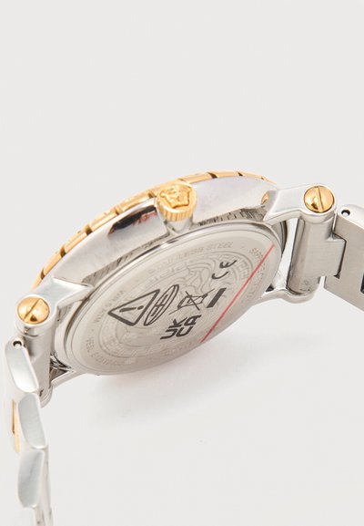 Πίσω μέρος ενός ρολογιού χειρός από ανοξείδωτο ατσάλι με χρυσές λεπτομέρειες, που διαθέτει κορώνα ρύθμισης και εγχάρακτα σύμβολα στο πίσω μέρος της κάσας.