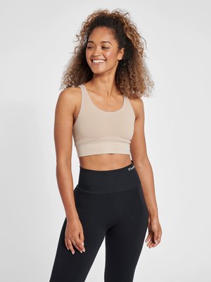 Smilende kvinde med krøllet hår iført beige sports-bh og sorte leggings med høj talje, stående mod en ensartet lys baggrund.