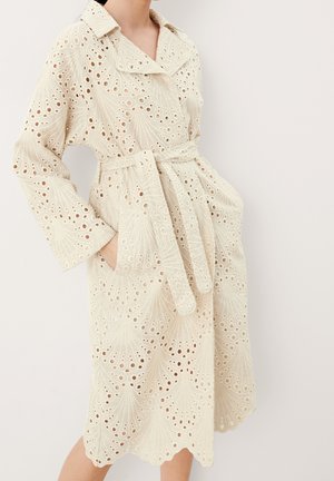 Femme portant un trench-coat en dentelle écru à œillets avec des bords festonnés et une ceinture nouée à la taille, debout les mains dans les poches.