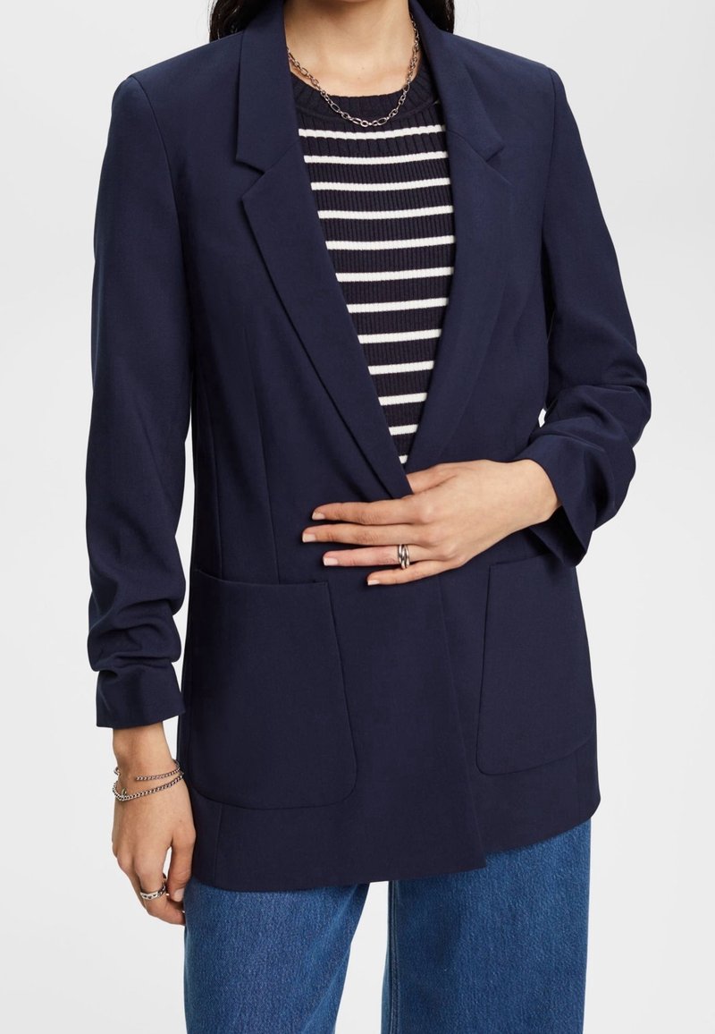 Marinblå blazer med två främre fickor, ensam knäppning och slagkrage, bärs över en marinblå och vit randig tröja.