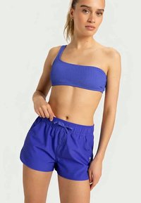 Μπλε ριχτό μπικίνι ενός ώμου, συνδυασμένο με μπλε κολυμβητικά σορτς με ελαστική μέση και κορδόνι. Απαλή υφή, άνετη εφαρμογή.