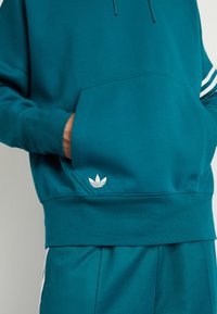 Felpa di cotone verde acqua con tasca frontale, orlo a coste e cappuccio con coulisse. Presenta tre strisce bianche sulle maniche e un piccolo logo.