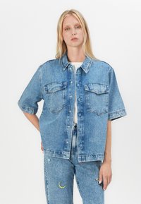 Cămașă din denim albastru deschis, cu mâneci scurte, buzunare frontale și închidere cu nasturi. Pur și simplu cu blugi albaștri asortați, cu broderie colorată.
