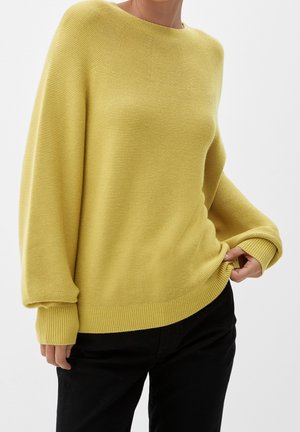 Vrouw draagt een loszittende gele gebreide trui met geribbelde manchetten en zoom, gecombineerd met een zwarte broek op een witte achtergrond.