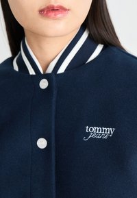 Jūrų tvido švarkas su baltu juostotu apykakle, išsiskiriantis sidabriniais snapelių mygtukais ir siuvinėtu "tommy jeans" logotipu krūtinėje.