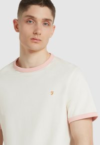 T-shirt color avorio con colletto e polsini a coste rosa. Presenta un piccolo logo arancione sulla zona sinistra del petto. Materiale in cotone morbido.