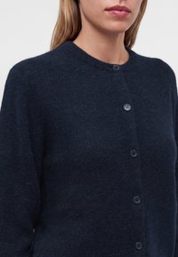 Femme portant un cardigan duveteux bleu marine foncé avec un col rond et cinq boutons visibles, visible des épaules jusqu'au milieu du torse.