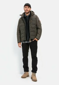 Oliven grøn quilted pufferjakke med hætte, sort skjorte, ternet flannel under, sorte bukser og beige snørebåndsstøvler.