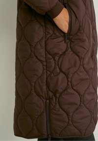 Hnědý prošívaný kabát s vlnovým vzorem, boční kapsou na zip, žebrovanými manžetami a rovnostranným střihem. Měkká textura a střední délka.