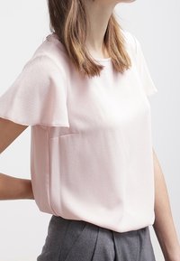 Ljust rosa kortärmad blus i ett texturerat tyg med rund halsringning och en ficka på sidan, i kombination med grå byxor.