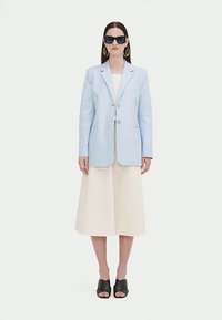 Woman wearing light blue blazer, cream midi skirt, black open-toe heels, large gold hoop earrings, and black sunglasses, standing against white background.