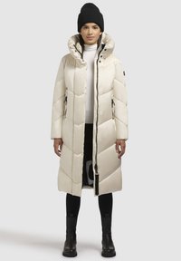 Puffjakke i creme med et diamantkviltmønster, høj krave, lynlås foran og sidelommer. Sammen med sorte støvler og en hue.