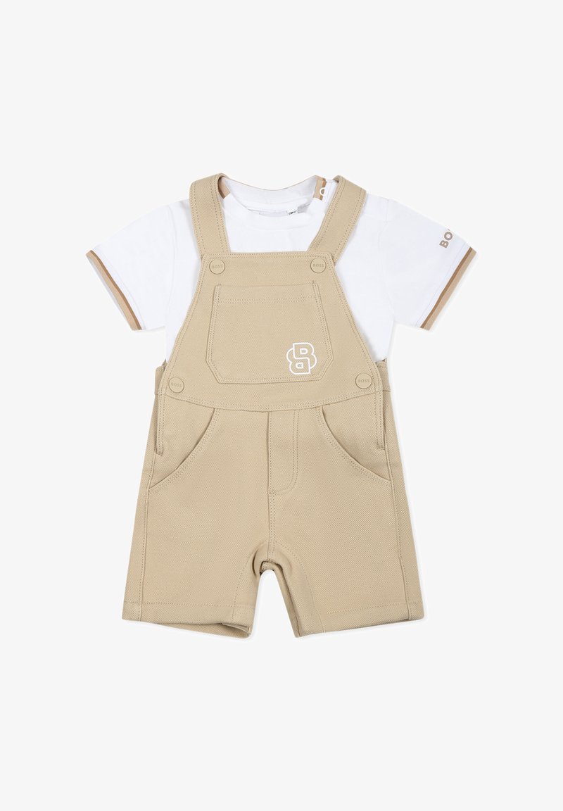 Salopette beige en tissu texturé, avec deux poches avant et un T-shirt blanc à manches courtes en dessous, agrémenté d'accents marron.