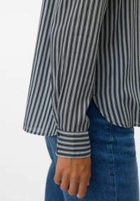 Højre arm iført en sort og hvid stribet langærmet skjorte med to knapper ved håndleddet, parret med blå denim jeans.