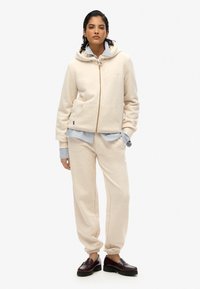 Felpa beige con zip, tasche e cappuccio con stringhe, abbinata a pantaloni jogger beige coordinati. Il modello indossa una maglietta a righe sotto.
