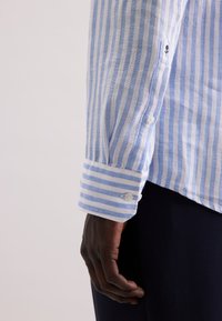 Camicia a righe blu chiaro e bianca a maniche lunghe con polsino abbottonato, realizzata in un morbido tessuto; presenta un piccolo ricamo floreale.