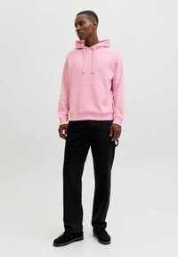 Vaaleanpunainen huppari, jossa on kiristysnauhat ja etutasku, yhdistetty mustiin housuihin ja mustiin kenkiin. Hupparissa on pieni kirjailtu yksityiskohta.