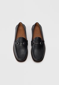 Mocasines de cuero negro con una textura suave, puntera redondeada y adornos de metal plateado decorativos en la parte frontal. Interior marrón y detalles de costura.