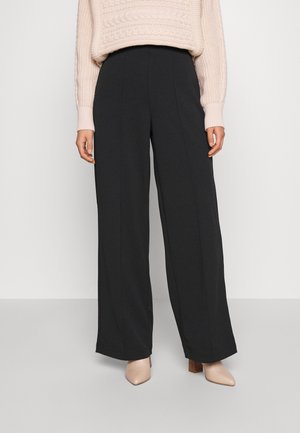 Vrouw die zwarte wide-leg broek, beige gebreide trui en beige schoenen met hak draagt, staand tegen een effen witte achtergrond.