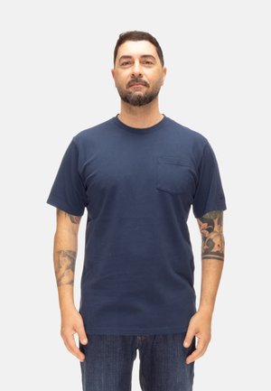 Man met kort donker haar en baard, gekleed in een marineblauw T-shirt en spijkerbroek, staand met zichtbare tatoeages op beide onderarmen tegen een effen achtergrond.