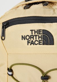 Rucsac bej cu fermoar negru, având logo-ul "The North Face" brodat în negru. Include un cordon elastic verde și un material texturat.