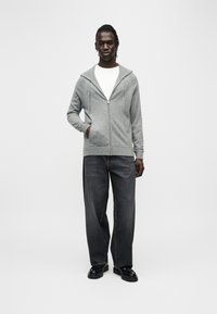 Sweat à capuche gris zippé avec capuche à cordon, associé à un jean ample de couleur foncée et des chaussures noires. Le tissu présente une finition texturée.