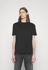 T-shirt noir à manches courtes en tissu doux, avec un col rond et un petit patch de marque sur la poitrine. Coupe décontractée, texture lisse.