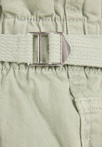 Tejido verde claro con una textura suave, que presenta una correa de hebilla plateada y una cinturilla elástica gathered para un ajuste ajustable.