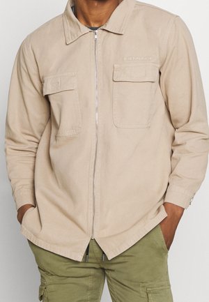 Uomo con camicia beige con zip e due tasche sul petto e pantaloni cargo verde oliva, mani nelle tasche.