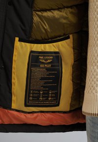 Zwart pufferjack interieur met een gouden voering en een gele mesh pocket met zorglabel en specificaties. Tekstdetails zichtbaar.