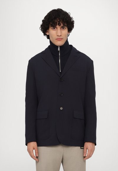 Blazer marine avec deux poches avant, fermeture à un bouton et une poche poitrine. En dessous, un haut noir à col montant avec fermeture éclair.