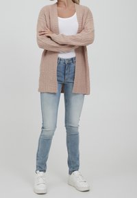 Ljusrosa stickad cardigan med öppen fram, tillsammans med ljusblå skinny jeans och vita sneakers. Cardiganen har ribbad textur och långa ärmar.
