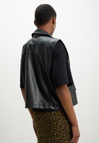 Schwarz-faux Lederweste mit Kragen, ärmellosem Design und Seitentaschen. Getragen über einem schwarzen T-Shirt und einem Leopardenmuster-Rock.