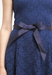 Robe en dentelle bleu marine avec un motif floral, dotée d'une large ceinture en satin nouée en nœud à la taille. Texture lisse et silhouette ajustée.