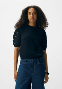 Femme aux cheveux bouclés portant un haut noir à manches courtes avec des découpes sur les manches et un jean en denim bleu foncé.
