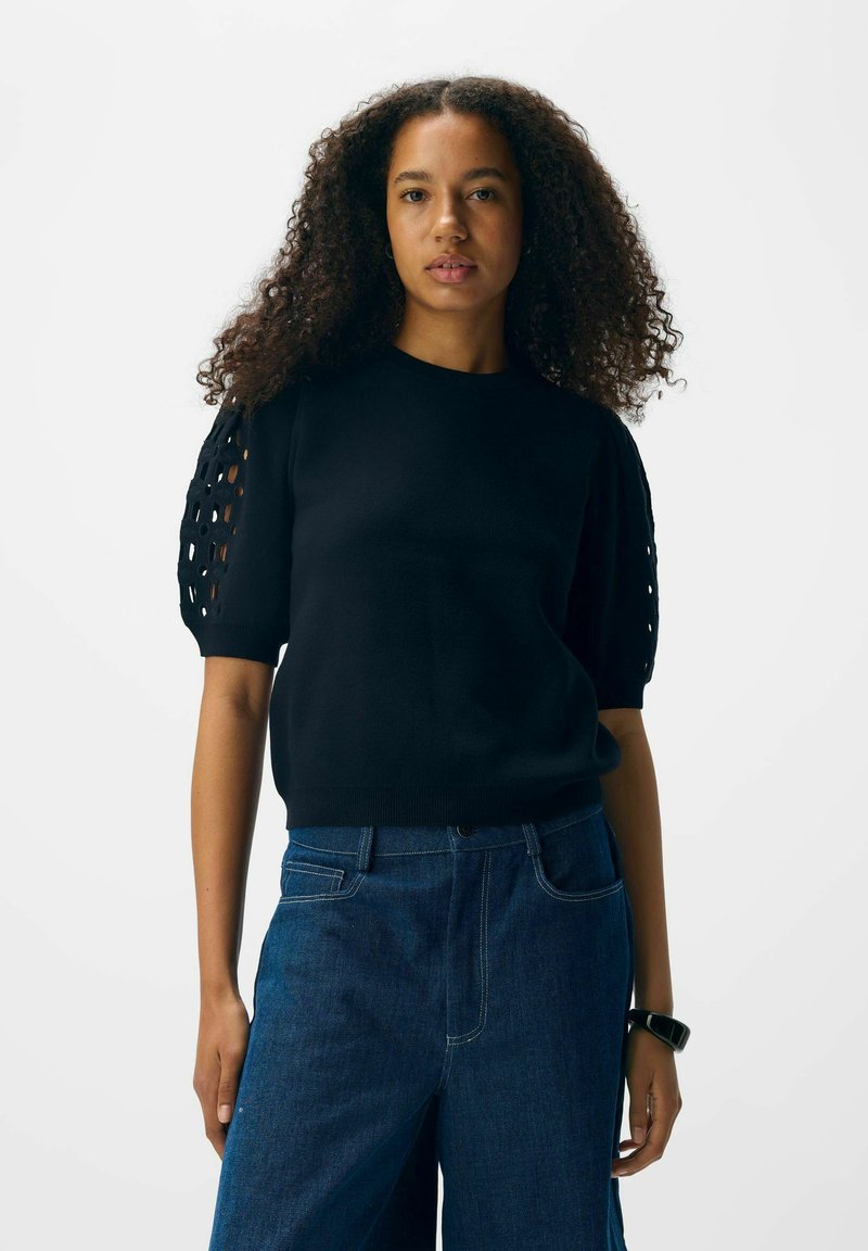 Femme aux cheveux bouclés portant un haut noir à manches courtes avec des découpes sur les manches et un jean en denim bleu foncé.
